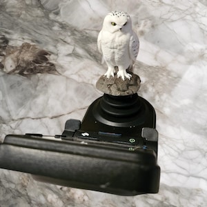May include: A black joystick controller featuring a white snowy owl figurine perched on a textured gray rock atop the handle. The base includes a black plastic housing with visible power and function buttons, showcasing a detailed avian sculpture.