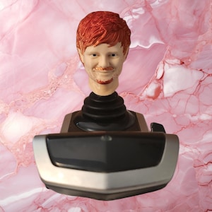 May include: A custom joystick handle featuring a sculpted, realistic head with textured ginger hair and a light skin tone, mounted on a black and silver gaming controller base with a black rubberized neck connector and a smooth, curved front panel.