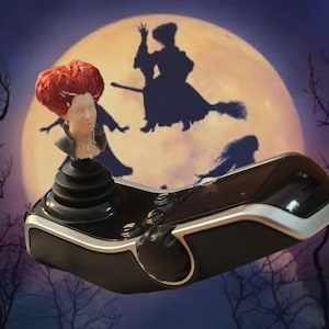 May include: A black gaming joystick controller featuring a sculpted figurine of a person with bright red hair and a dark collar attached to the top of the stick. The controller has a smooth, curved black body with silver trim accents along the edges.