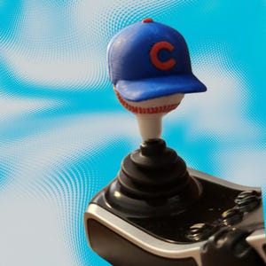 May include: A blue baseball cap with a red letter C on the front, resting on top of a white baseball with red stitching. This baseball-themed topper is attached to a black joystick controller base with a ribbed rubber shaft and silver accents.
