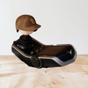 May include: A black and silver handheld video game controller featuring a baseball-themed joystick topper. The topper consists of a white baseball wearing a brown San Diego Padres baseball helmet with the gold SD logo clearly visible on the front.