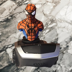 May include: A Spider-Man bust figurine featuring a textured orange and blue suit with a black spider emblem on the chest, mounted atop a black and silver gaming joystick base with a smooth, curved front panel and a circular speaker grille.