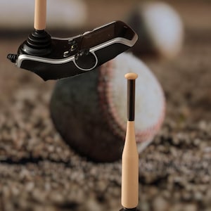 May include: A black and silver joystick with a baseball bat-shaped handle. The handle is light brown with a dark brown top. A baseball is in the background, resting on a dirt surface. The joystick has buttons and a cord.