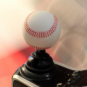 May include: A baseball-themed joystick with a white baseball featuring red stitching atop a black base. The joystick has a sleek, modern design with a black and silver color scheme, suggesting a gaming or sports-related product.