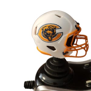 May include: A white miniature American football helmet with an orange face mask and the Chicago Bears logo. The logo features a bear head inside a navy blue C with orange and navy blue accents. The helmet is mounted on a black base.