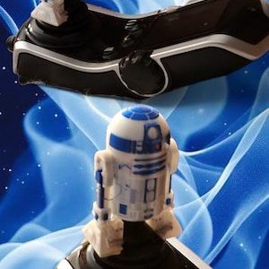 May include: A black and silver joystick controller with a white and blue R2-D2 figurine on top. The controller has buttons and a sleek design. The background is a blue abstract design.