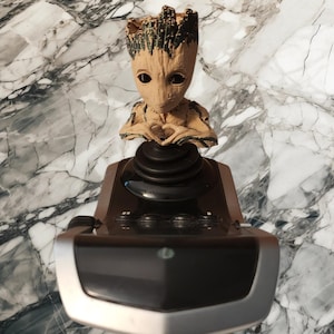 May include: A tan and dark brown tree-like character figurine with large black eyes and hands forming a heart shape, mounted atop a black and silver joystick controller featuring multiple circular buttons and a smooth, rounded base design.