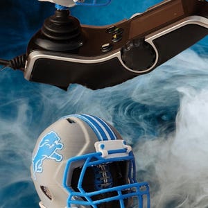May include: Two miniature Detroit Lions football helmets, one atop a black joystick and the other below. The helmets are gray with blue stripes and a blue lion logo. The background is a blue gradient with wisps of white.