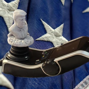 May include: A gray bust of a man atop a black and brown device, set against a blue background with white stars. The device has buttons and a joystick. The background resembles a flag.