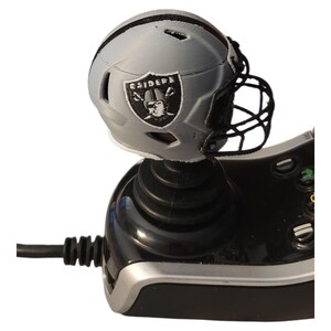 May include: A black video game controller joystick topped with a miniature silver and black football helmet. The helmet features a black face mask and a Raiders team logo on the side, which includes the text Raiders and a pirate illustration.
