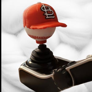May include: A black gear shift knob featuring a white baseball topper with red stitching. An orange baseball cap with a white and black St. Louis Cardinals logo is positioned on top of the baseball. The base is black with a ribbed rubber boot.