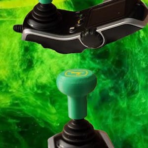May include: Two black and silver joysticks with green tops and the John Deere logo. The joysticks have a black base and are set against a green and yellow background.