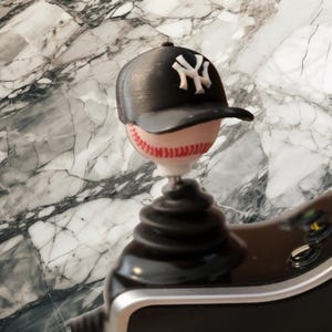 May include: A black baseball cap with a white NY logo sits atop a white baseball with red stitching. The baseball is attached to a black, ribbed, conical gear shift knob base with a silver trim, designed for use as a vehicle interior accessory.