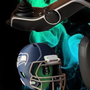 May include: Miniature football helmets in navy blue with the Seattle Seahawks logo, mounted on black joystick controllers. The helmets have gray face masks. The background is black with teal and green smoke.