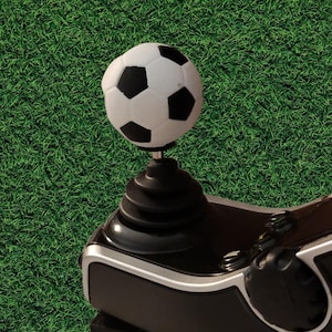 May include: A black and white soccer ball shaped gear shift knob attached to a black, ribbed, conical base. The gear shifter features a spherical top with a classic pentagonal pattern and is mounted on a black and silver mechanical control unit.