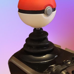 May include: A red and white Pokeball-themed joystick topper. The topper is mounted on a black base, with a black rubber boot. The design is reminiscent of the popular animated series.