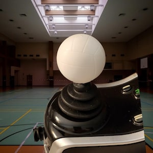 May include: A black and silver joystick controller featuring a white, spherical volleyball-shaped topper. The controller has a tiered black rubber base and several small, circular buttons integrated into the glossy black surface of the device.