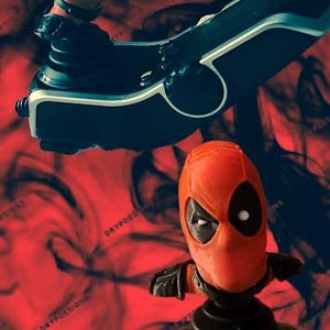 May include: Two Deadpool figures, one seated on a black and white vehicle, the other standing on a black base. The figures are red and black, with white eyes. The background is a red and black abstract design.