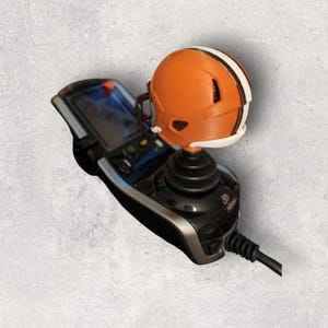 May include: An orange and white miniature football helmet rests on top of a black joystick controller. The controller has a screen and buttons. The helmet is a replica of a Cleveland Browns helmet.