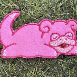 May include: Two identical pink embroidered patches featuring a stylized creature with a curled tail, rounded ears, and a wide, smiling mouth. The patches have a felt-like texture and are outlined with a darker pink thread, showcasing a flat design.