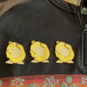 May include: Black quarter-zip sweatshirt with three yellow Psyduck patches. The sweatshirt features a colorful patterned trim around the collar and a decorative band with desert landscape imagery. The zipper is black.
