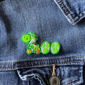 May include: A green embroidered patch featuring a cartoon dinosaur character sitting next to two light green spotted eggs, attached to the front of a blue denim jacket with metal buttons and visible stitching along the pockets and seams.