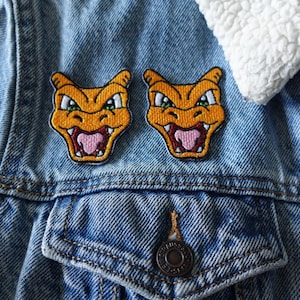 May include: Two embroidered orange Charizard patches with green eyes and pink tongues are sewn onto a light blue denim jacket. The jacket has a white, fluffy collar and a metal button closure.