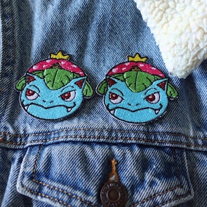 May include: Two embroidered patches featuring a blue creature with red eyes, green leaves, and a pink and yellow crown. The patches are affixed to a denim jacket with a white, fluffy collar.