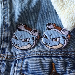 May include: Two embroidered patches featuring a blue cartoon character with brown and white accents. The patches are sewn onto a denim jacket with a cream-colored, fuzzy collar. The jacket is a light blue wash.