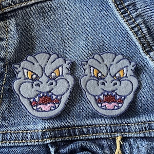 May include: Two embroidered patches featuring the head of a grey monster with yellow eyes, a dark blue border, and an open mouth showing sharp teeth and a red tongue. The patches are attached to a blue denim fabric with visible stitching and seams.