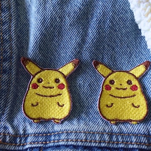 May include: Two yellow Pikachu embroidered patches with brown outlines and red cheeks are sewn onto a blue denim jacket. The patches are side by side. A white, fluffy fabric is in the upper right corner.