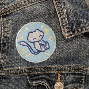 May include: A round, light blue embroidered patch featuring a cartoon creature with a long tail, sitting with its legs crossed. The patch is surrounded by a light blue border and yellow star accents. The patch is sewn onto a denim jacket.