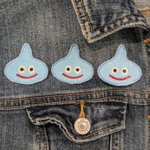 May include: Three light blue, droplet-shaped patches with smiling faces are affixed to a denim jacket. Each patch has a red smiling mouth, black eyes, and a light blue outline. The patches are evenly spaced.