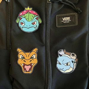 May include: Black backpack with four embroidered patches of cartoon characters. The patches are colorful and feature a blue, green, and red character, an orange and yellow character, and a blue and white character. The backpack has a Vans label.