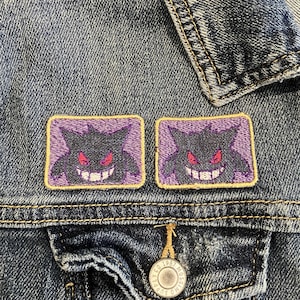May include: Close-up of a denim jacket with two embroidered patches. Each patch features a purple cartoon ghost with a sinister grin and red eyes, set against a lavender background. The patches are outlined in yellow thread.