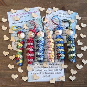 Könnte beinhalten: Handgemachte Holzpuppen mit gehäkelten Körpern in verschiedenen Farben, darunter Rot, Blau und Gelb. Jede Puppe hat ein lächelndes Gesicht und einen kleinen Hut. Die Puppen sind mit hölzernen Herz-Ausschnitten und Karten mit deutschem Text angeordnet.