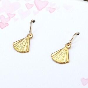 May include: A pair of gold-toned earrings with a fan-shaped design. The earrings are on a white background with pink hearts.