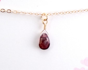 May include: A delicate gold chain necklace with a small, teardrop-shaped, dark red gemstone pendant.