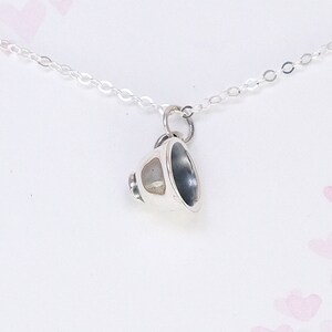May include: A silver necklace with a small, delicate teacup charm. The necklace is on a white background with a pattern of pink hearts.