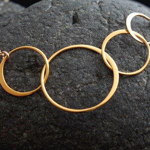 May include: A gold necklace with five interlocking circles of varying sizes. The necklace is on a dark gray textured surface.