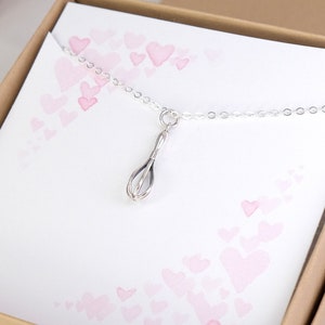Puede incluir: Un collar de plata con un dije de batidor. El collar est&aacute; en una caja con un fondo rosa y blanco con corazones.