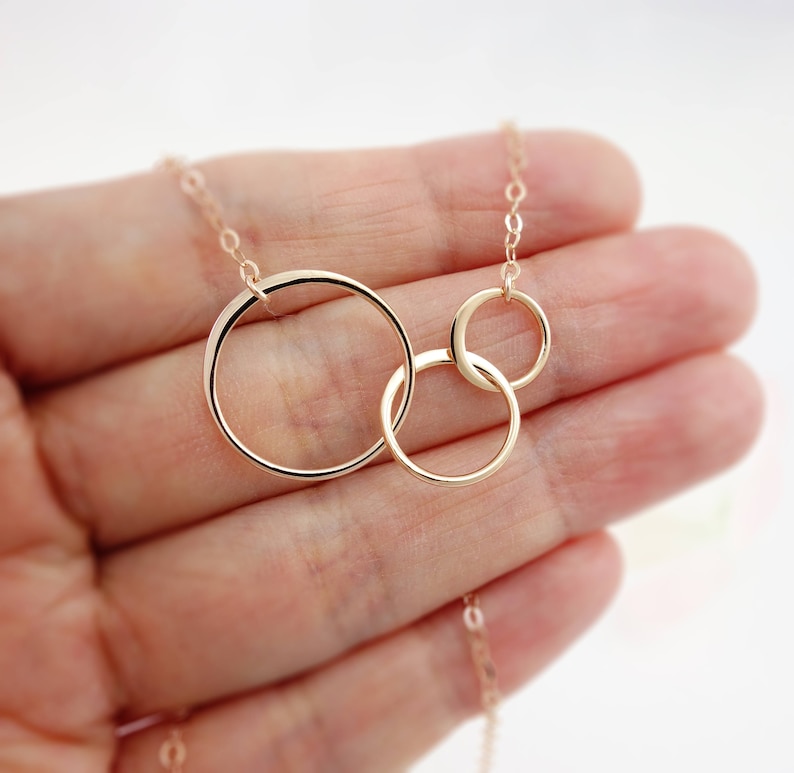 Peut inclure: Un collier en or avec trois cercles entrelac&eacute;s. Les cercles sont de tailles diff&eacute;rentes et sont reli&eacute;s les uns aux autres. Le collier est sur une main.