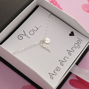 May include: A silver necklace with a small angel wing charm and a personalized initial charm. The necklace is in a white box with a card that reads "You... Are An Angel".