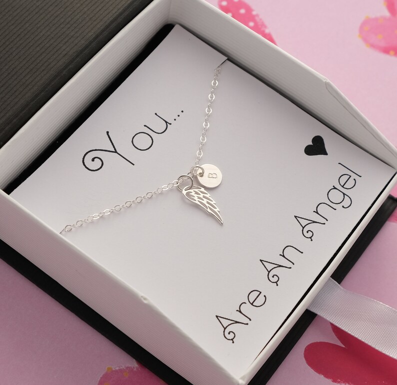 May include: A silver necklace with a small angel wing charm and a personalized initial charm. The necklace is in a white box with a card that reads "You... Are An Angel".