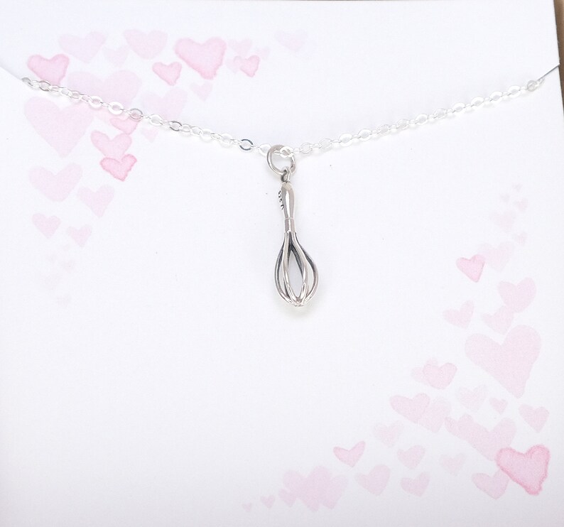 Puede incluir: Un collar de plata con un peque&ntilde;o dije de batidor. El collar est&aacute; sobre un fondo blanco con corazones rosas de acuarela.