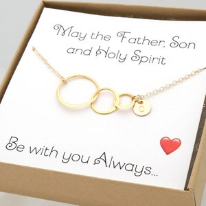 May include: Gold-colored necklace with three interlocking circles and a small disc pendant with the letter "C". The necklace is presented on a card with the text "May the Father, Son and Holy Spirit Be with you Always..." and a red heart.