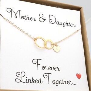 Pode incluir: Colar de ouro com dois c&iacute;rculos entrela&ccedil;ados e um pequeno pingente com a letra 'A'. O colar &eacute; apresentado em um cart&atilde;o branco com o texto 'M&atilde;e e Filha Unidas para Sempre...' e um cora&ccedil;&atilde;o vermelho.