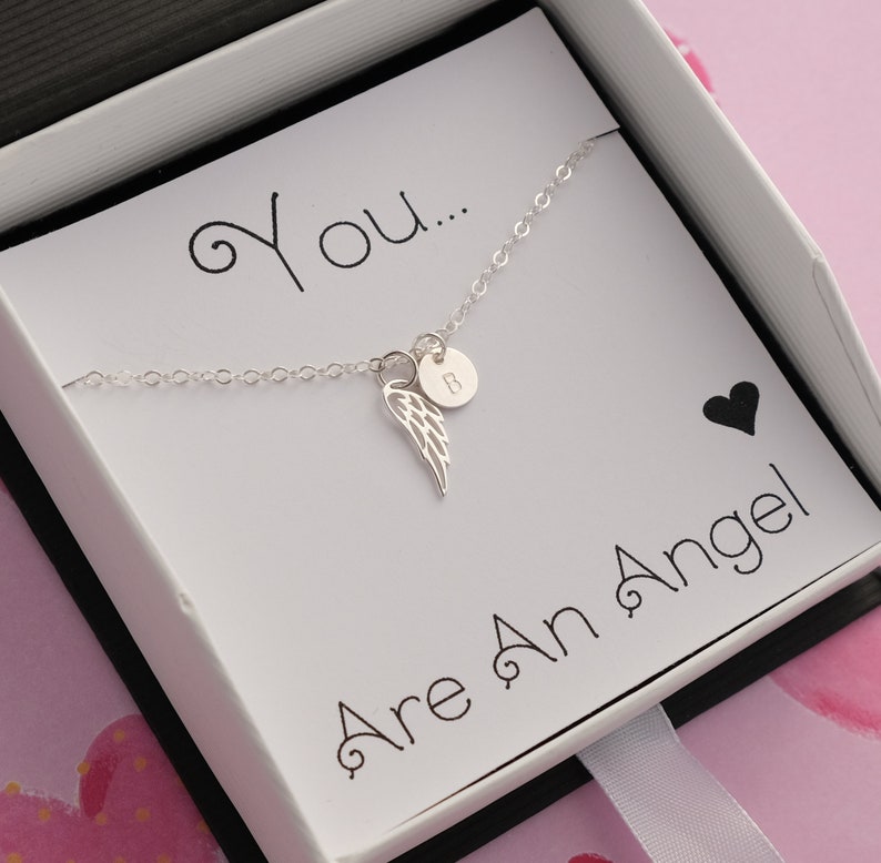 May include: A silver necklace with a small angel wing charm and a personalized initial disc. The necklace is in a white box with the text "You... Are An Angel!"