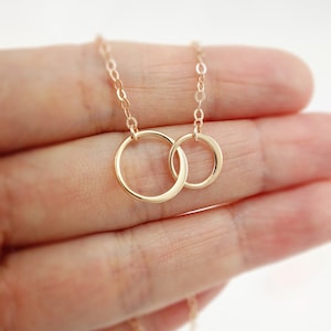 Pode incluir: Um colar de ouro com um pingente de dois c&iacute;rculos entrela&ccedil;ados. O colar est&aacute; em uma m&atilde;o.