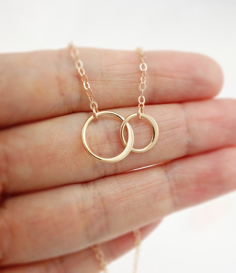 Pode incluir: Um colar de ouro com um pingente de dois c&iacute;rculos entrela&ccedil;ados. O colar est&aacute; em uma m&atilde;o.